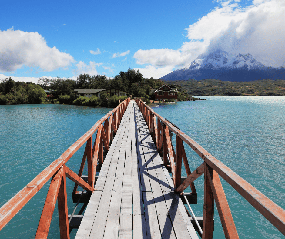 A wooden bridge over water with a house and mountains in the background

Description automatically generated