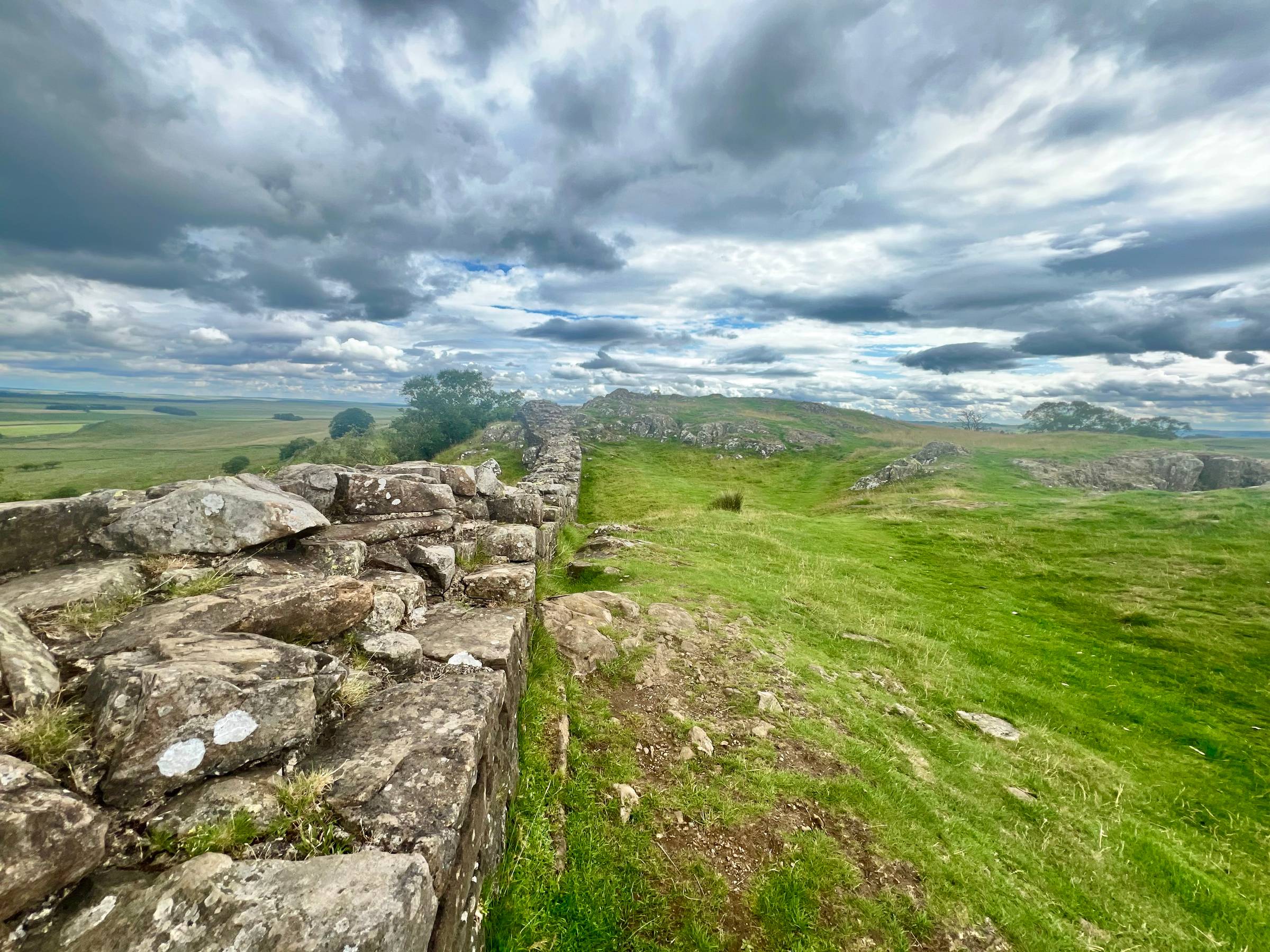 A photograph of a green field of grass with a few large rocks and trees in the background. There is an old stone wall which comes from the bottom left of the image, ending at the horizon line, slightly off to the left. The horizon line is just above the m