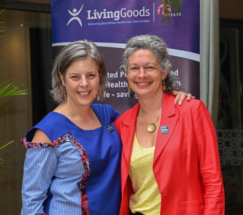 Portrait of Liz Jarman with her left arm on the shoulders of another woman. Both are smiling and standing, the photo is from the waist up. Liz is wearing a Blue asymmetric long sleeve top. The one side has a small cut out of the shoulder. The same side ha