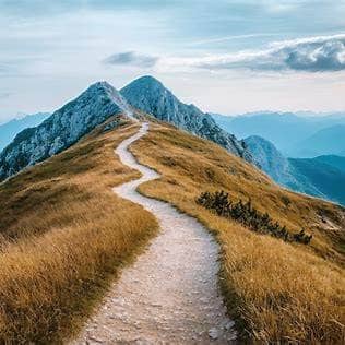 An image on top of mountains, with a winding dirt path down the middle which leads to more mountains in the distance. There is dry or straw colour grass on either side of the path. The mountains int he background and the top third of the image is shades o