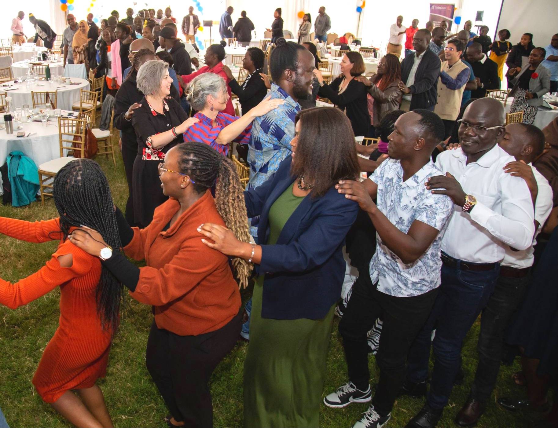 An photo of a group of racially diverse people in a winding congo line. The setting appears to be a wedding with round table which the people are winding through. There image has been take from slight above angle, giving a tilted, slight birds eye view of