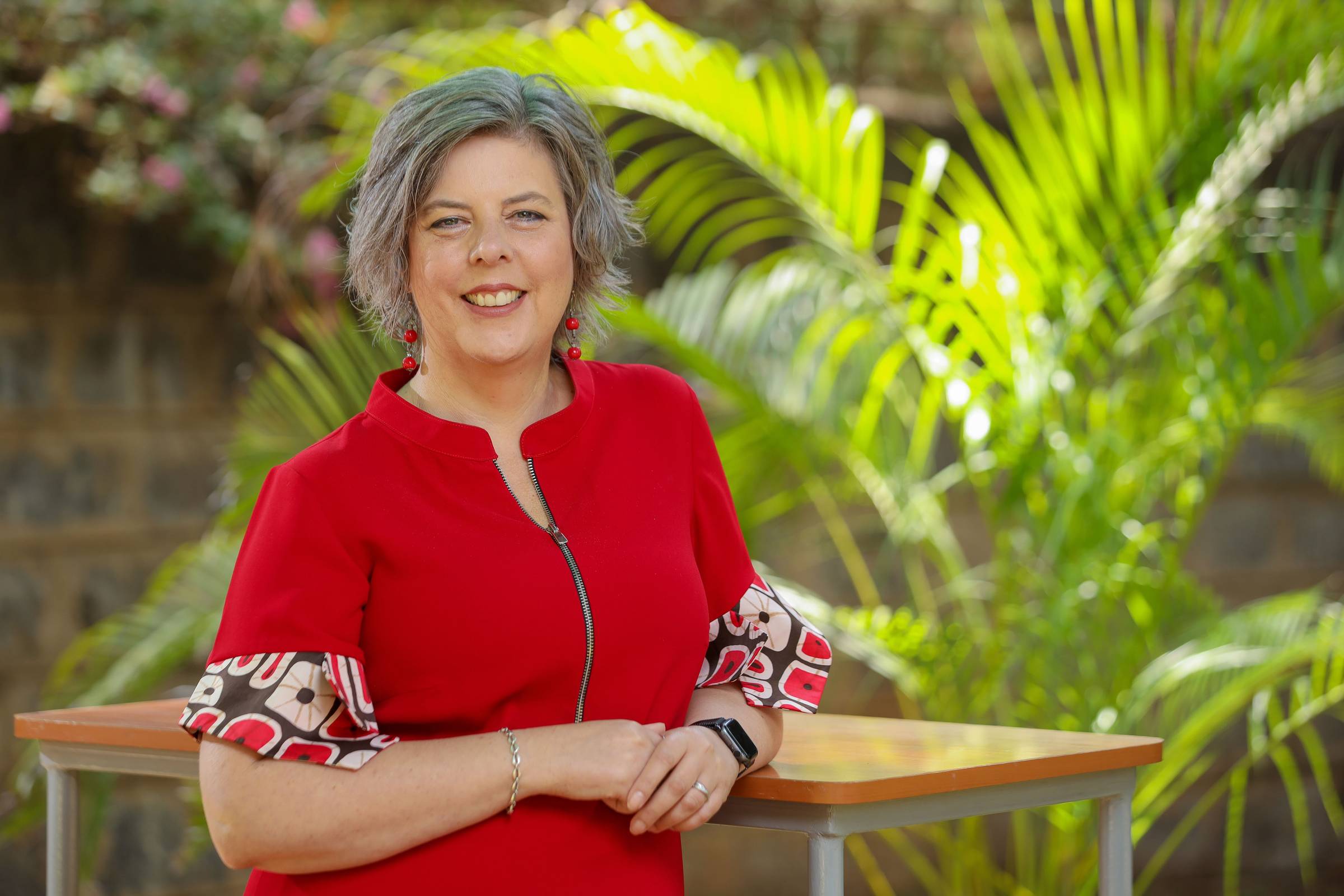 Portrait picture of Liz Jarman from the waist up, smiling. She is standing and resting her left arm on a table behind her. She is wearing a red dress short sleeves. The Sleeves have cuffs which are adored with square patterns, cream, red and brown coloure
