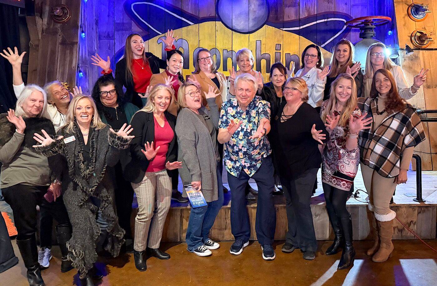 A group photo taken in front of a stage inside a restaurant, showing 18 people in various jazz hands pose. 