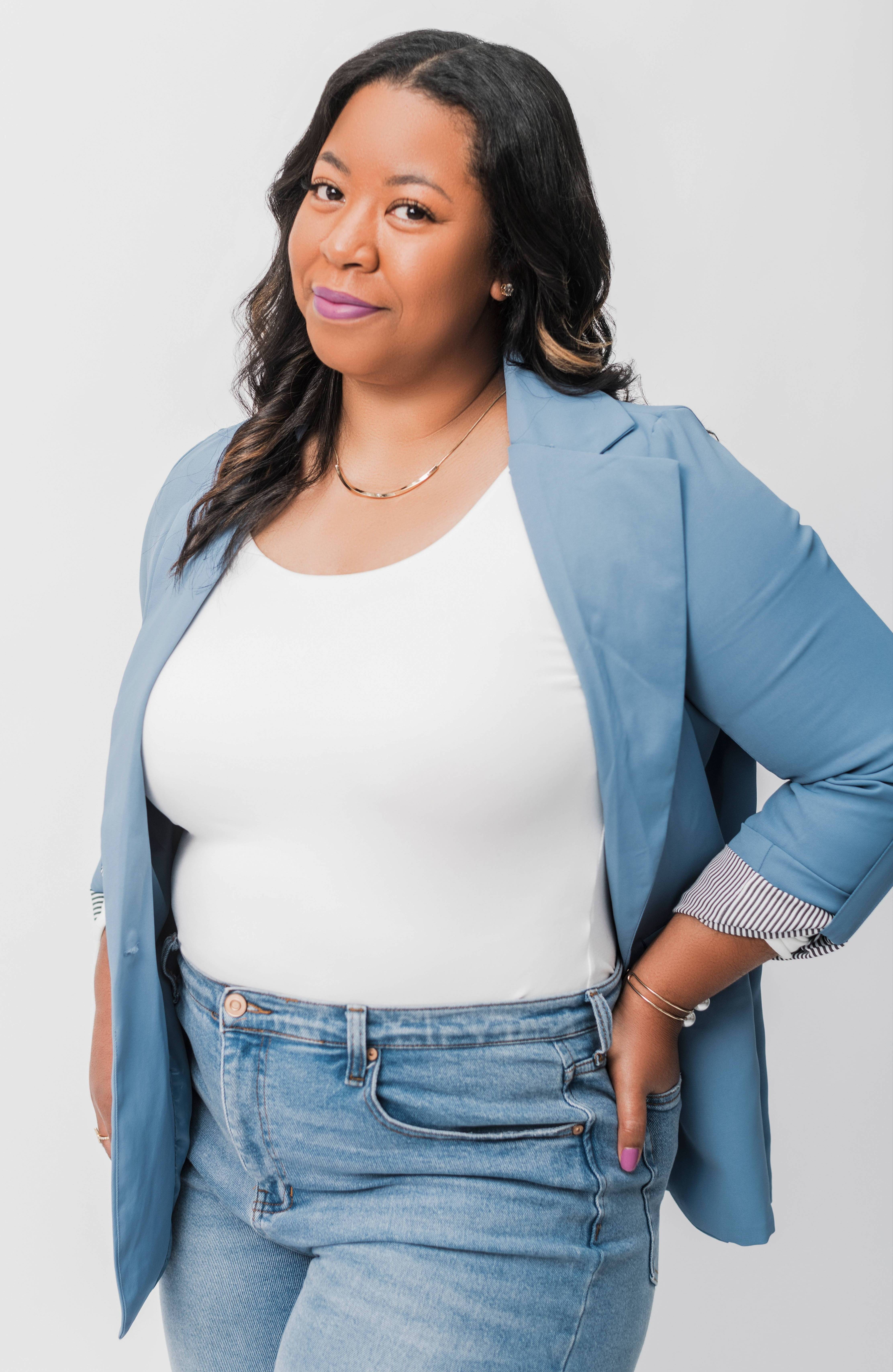 A woman wearing a blue blazer over a white top and jeans poses against a plain white background. She has long, wavy dark hair and is smiling softly, with her left hand in her pocket.