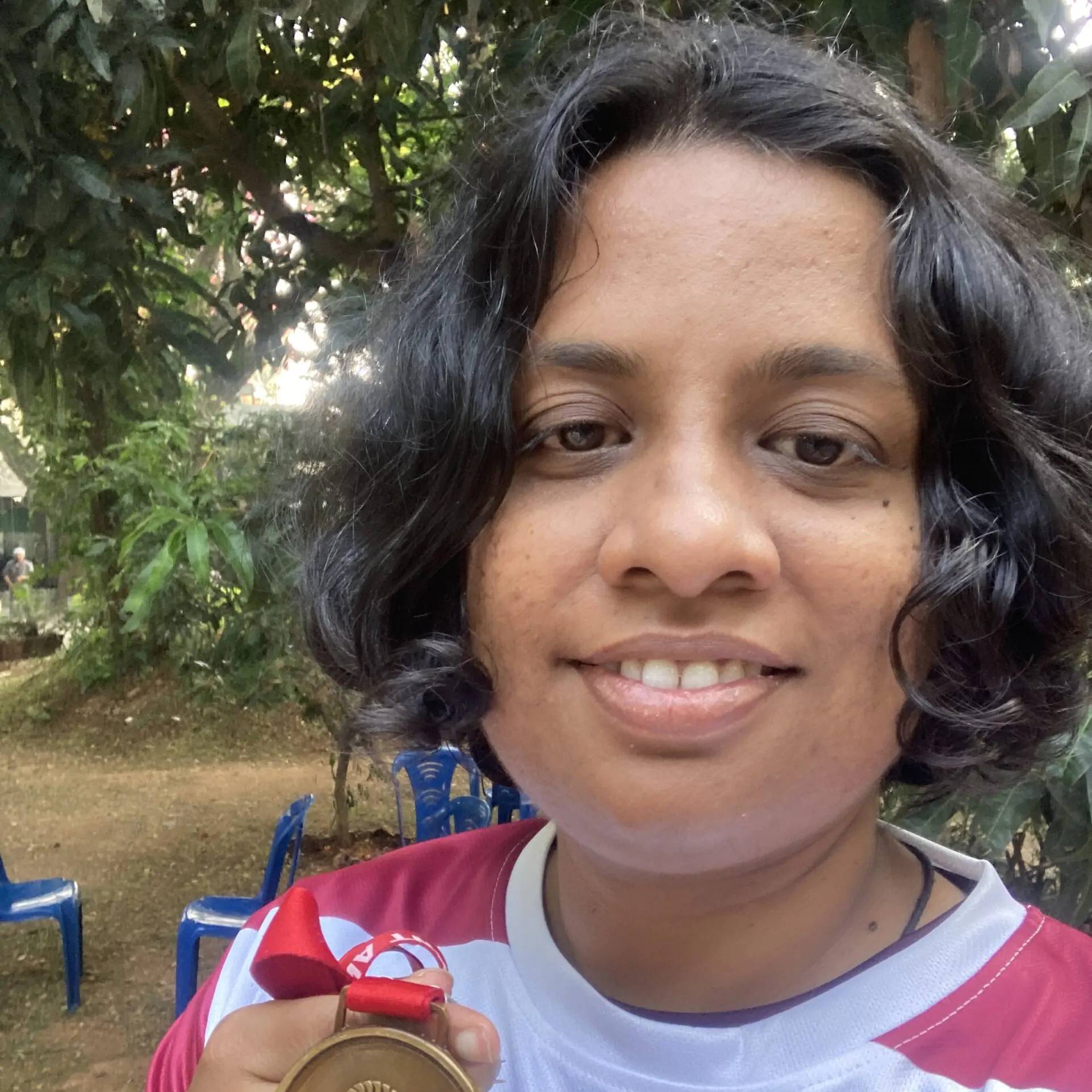 A person with short curly hair smiles at the camera while holding a medal with a red ribbon. They are wearing a sports jersey with white and maroon colors. In the background, there are green trees and several blue plastic chairs outdoors.