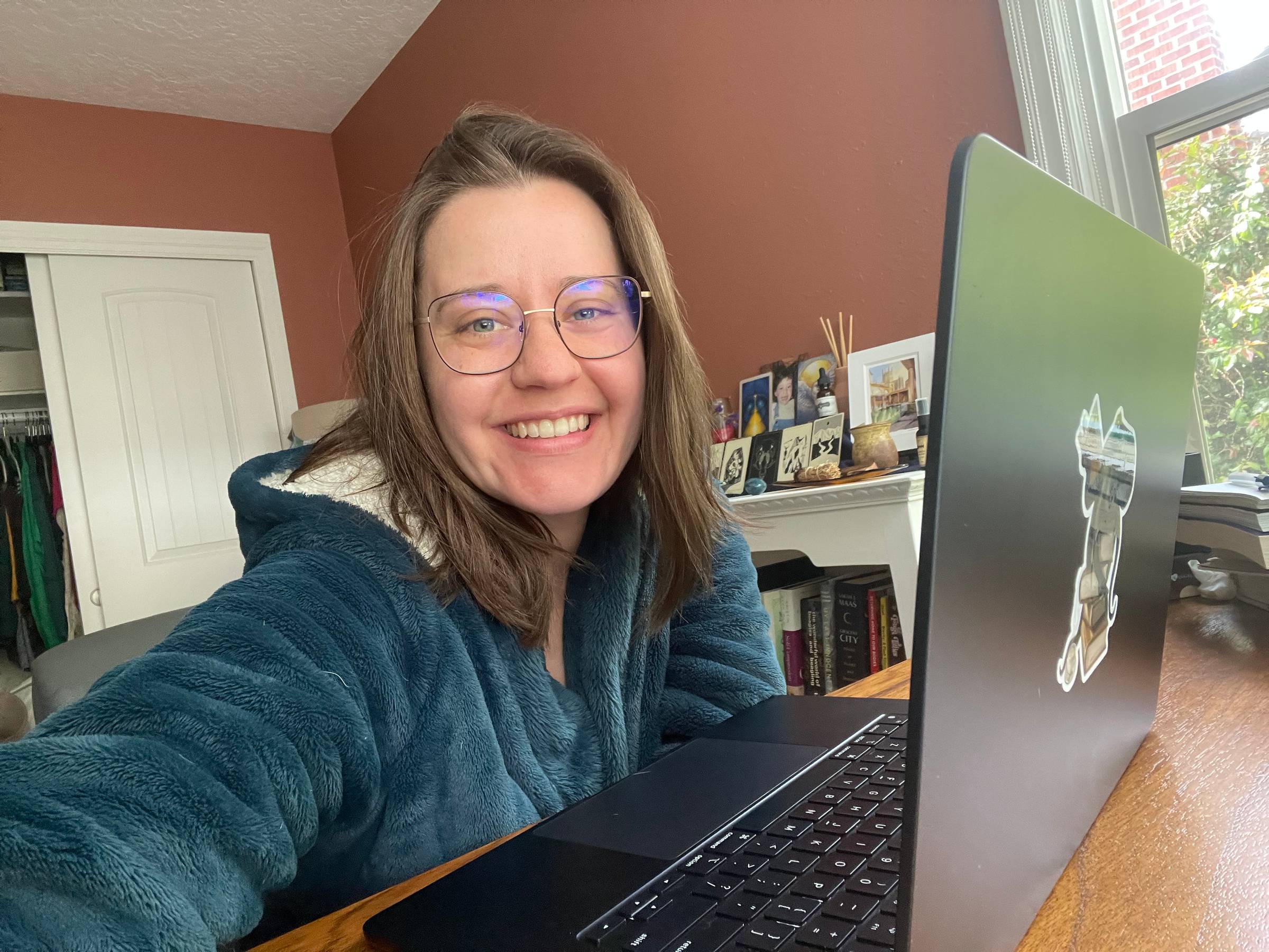 Image is a selfie taken by Maegan, a white woman with glasses and medium length brown hair, sitting in front of her laptop in a teal bathrobe, smiling at the camera.
