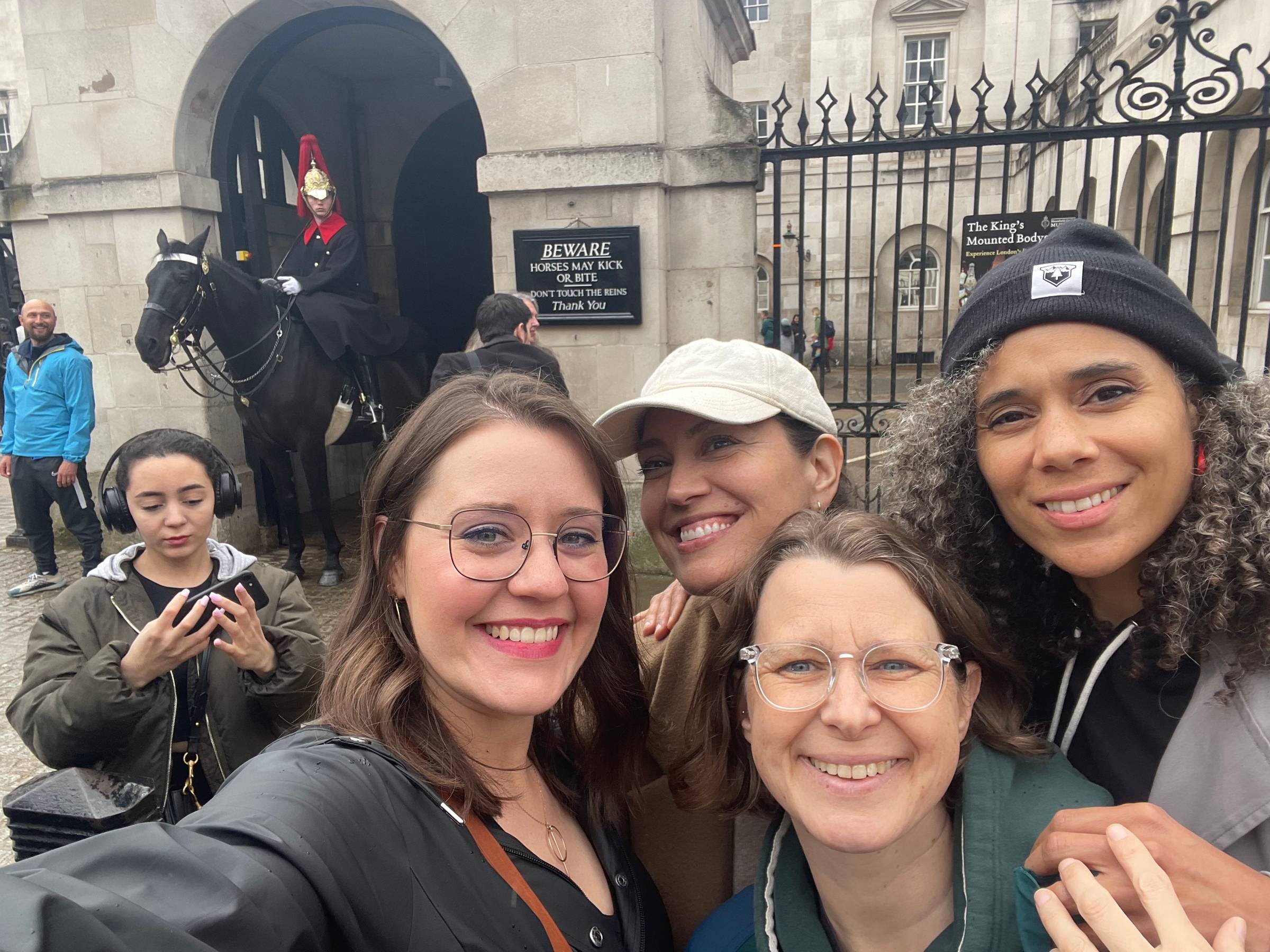 Image is a selfie of Maegan, Linzy, Annie, and Tiffany standing outside a gated palace, with a guard on a horse in the background.