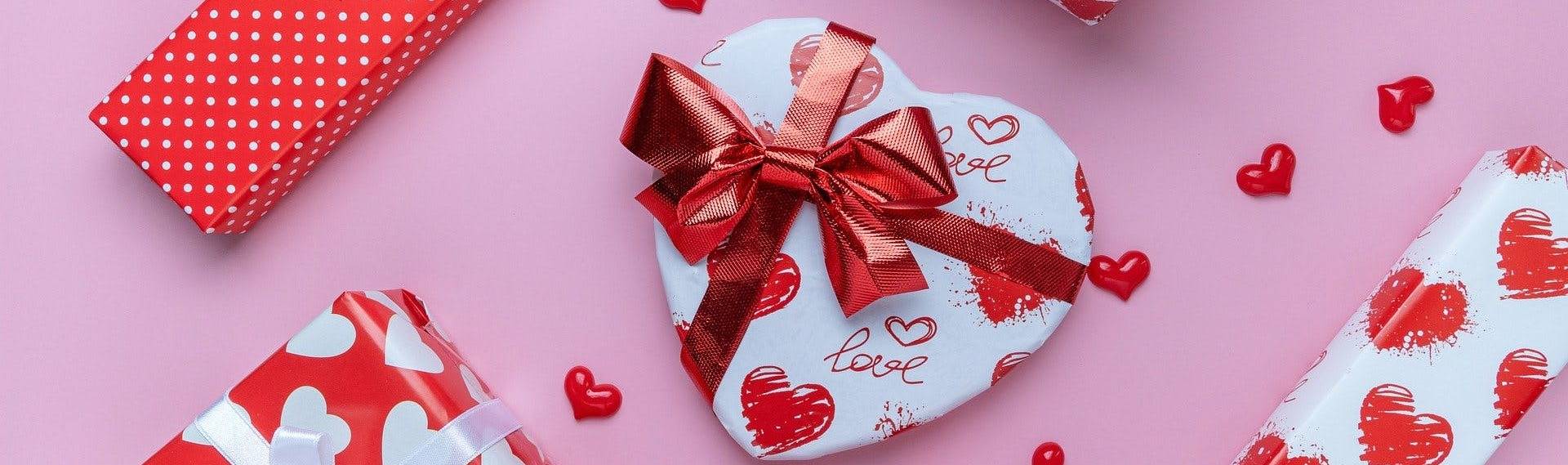 Assortment of gifts wrapped in Valentine's Day-themed read and white paper against a pink background.