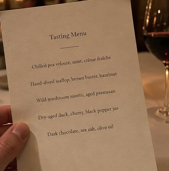 A hand holding a single-page tasting menu at a candlelit fine-dining table: five short courses on cream paper