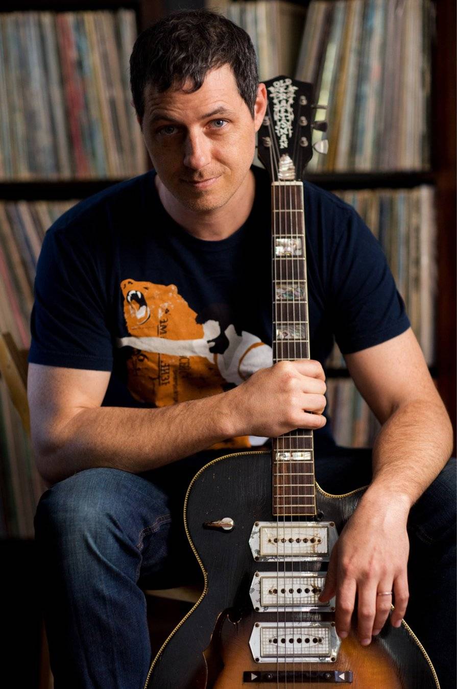 Lee Barbour seated with his archtop guitar in front of shelves of vinyl records