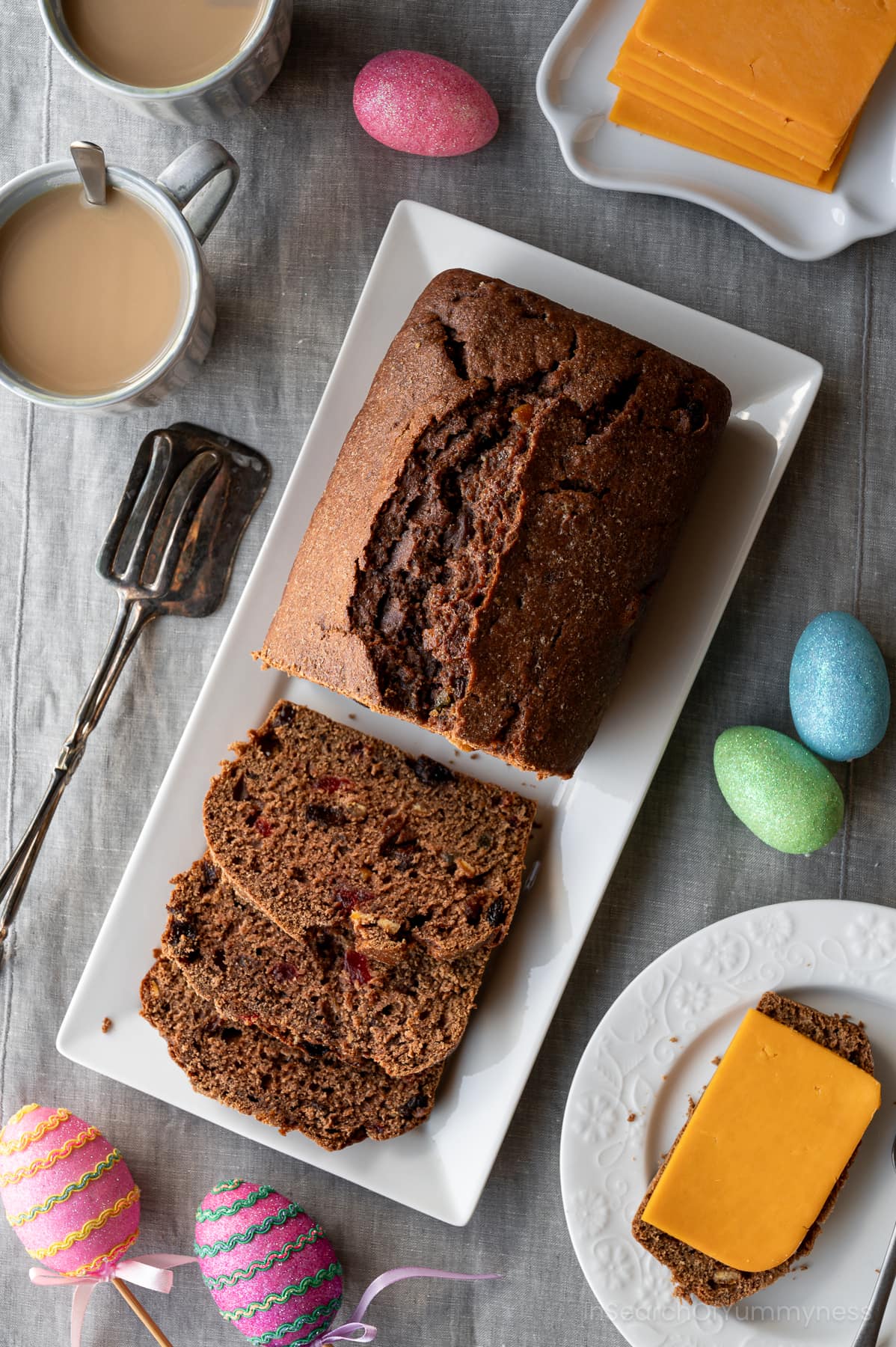A Jamaican Easter Bun loaf on a white platter next to cups of tea and Easter decorations.