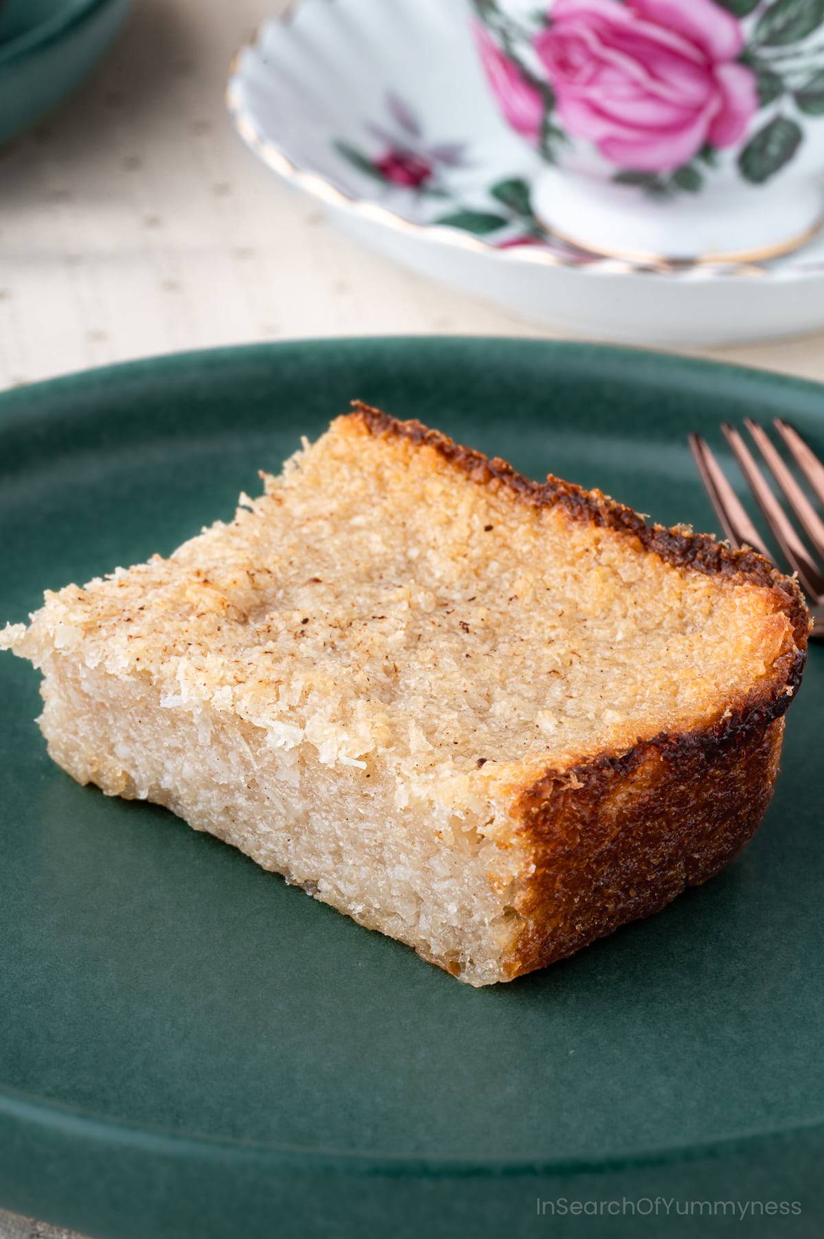 A slice of cassava pone on a green plate next to a fork and a tea cup.