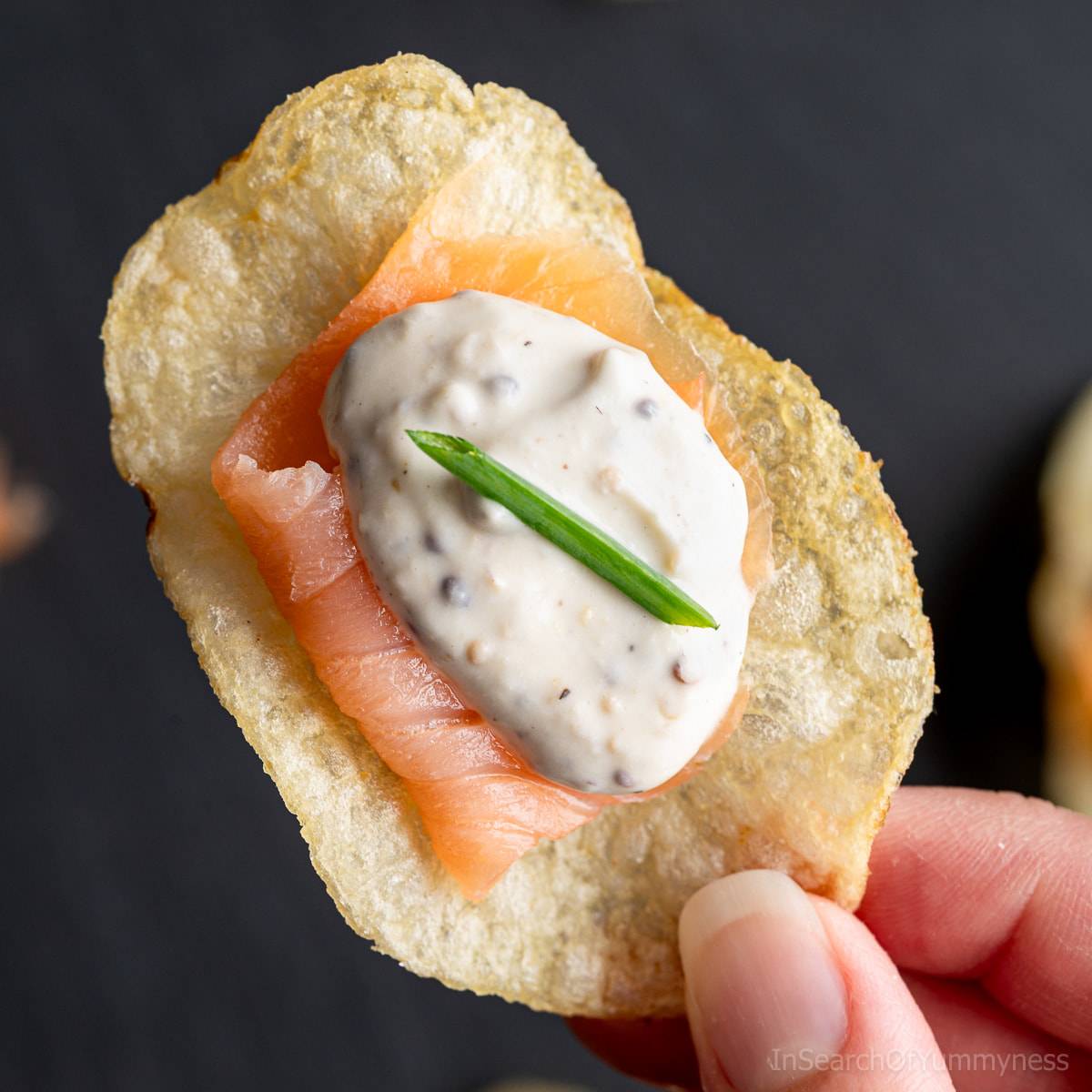 A hand holding a crisp plain potato chip topped with a piece of smoked salmon, a dollop of creme fraiche, and a fresh chive.