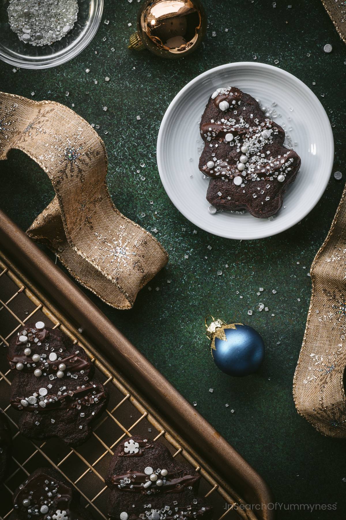 A small white plate with a tree-shaped chocolate shortbread cookies decorated with sprinkles next to a tray with more cookies.