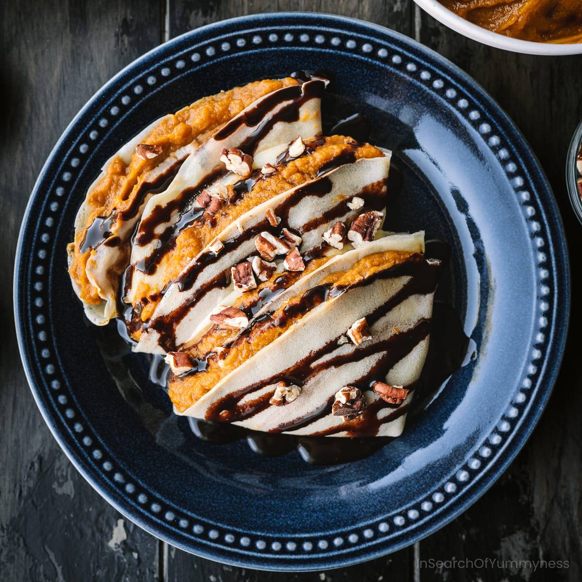 Three pumpkin crepes on a blue plate, topped with chocolate sauce and pecans.