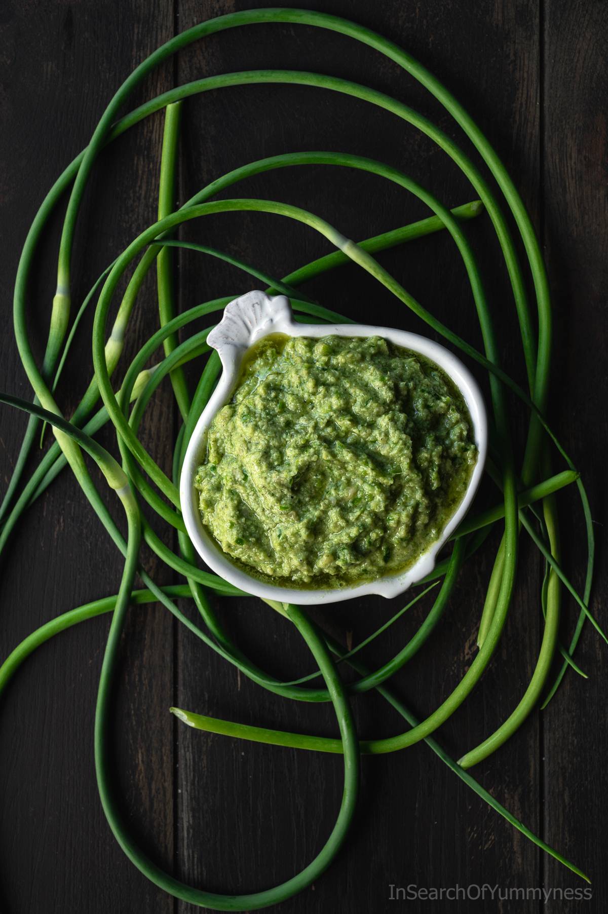 A garlic-shaped bowl full of garlic scape pesto, surrounded by fresh curly garlic scapes.
