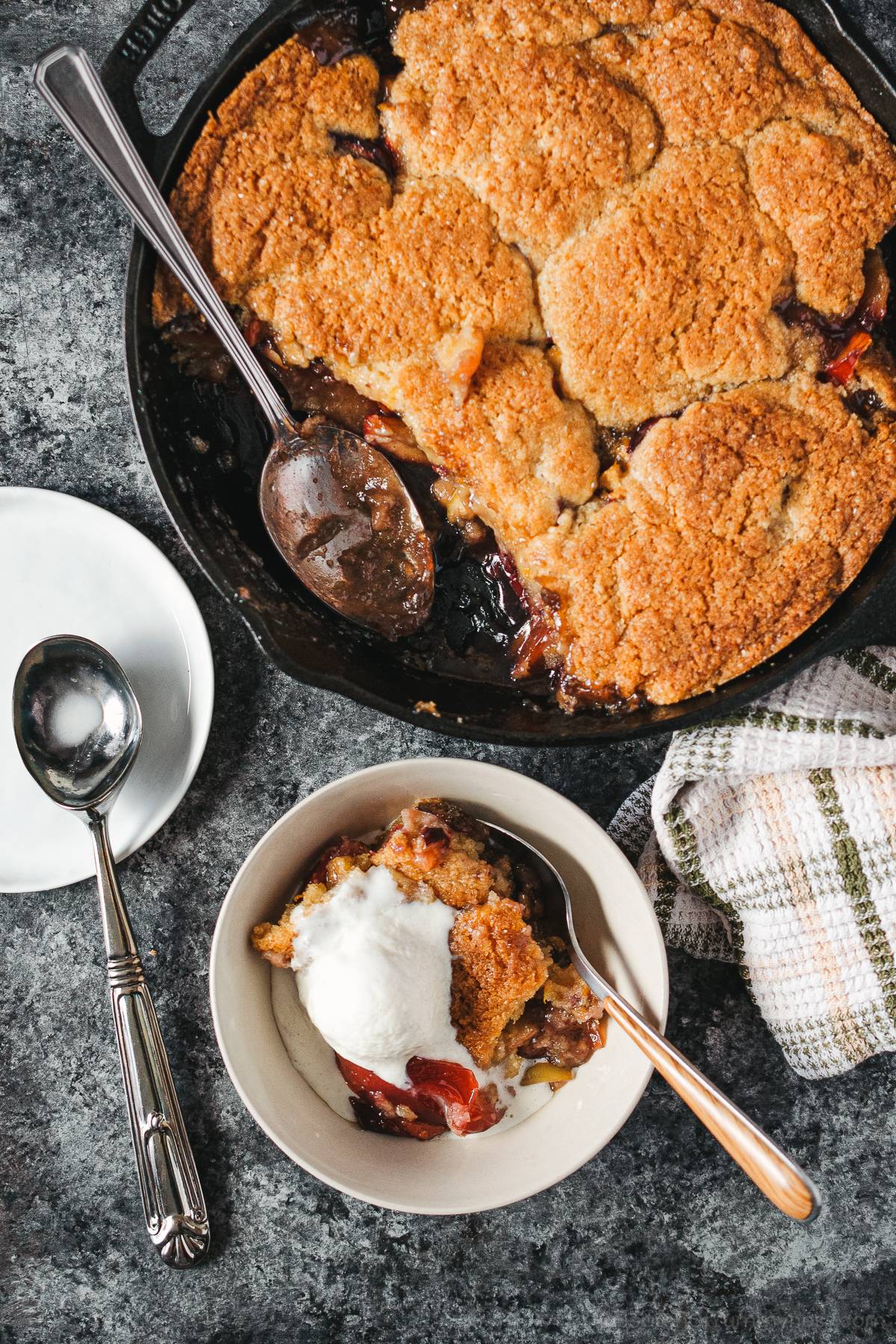 A cast iron pan with freshly baked plum cobbler, next to a bowl of cobbler topped with vanilla ice cream.