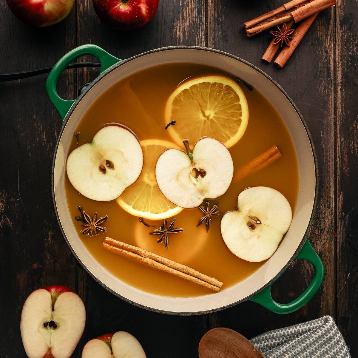 A green Le Crueset french oven full of warm spiced apple cider with slices of apples and oranges floating in it.