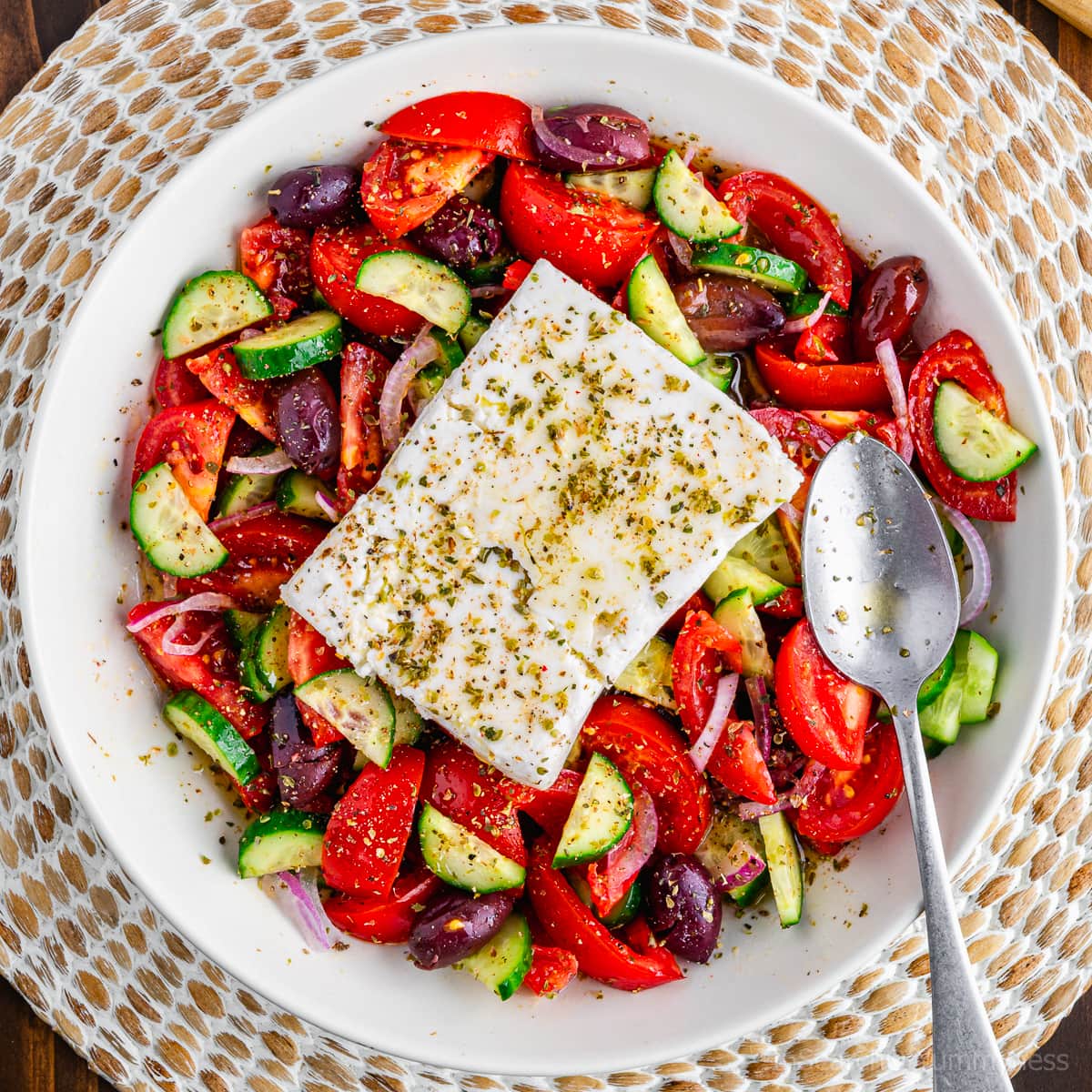 A white bowl full of Greek salad topped with a block of feta cheese.