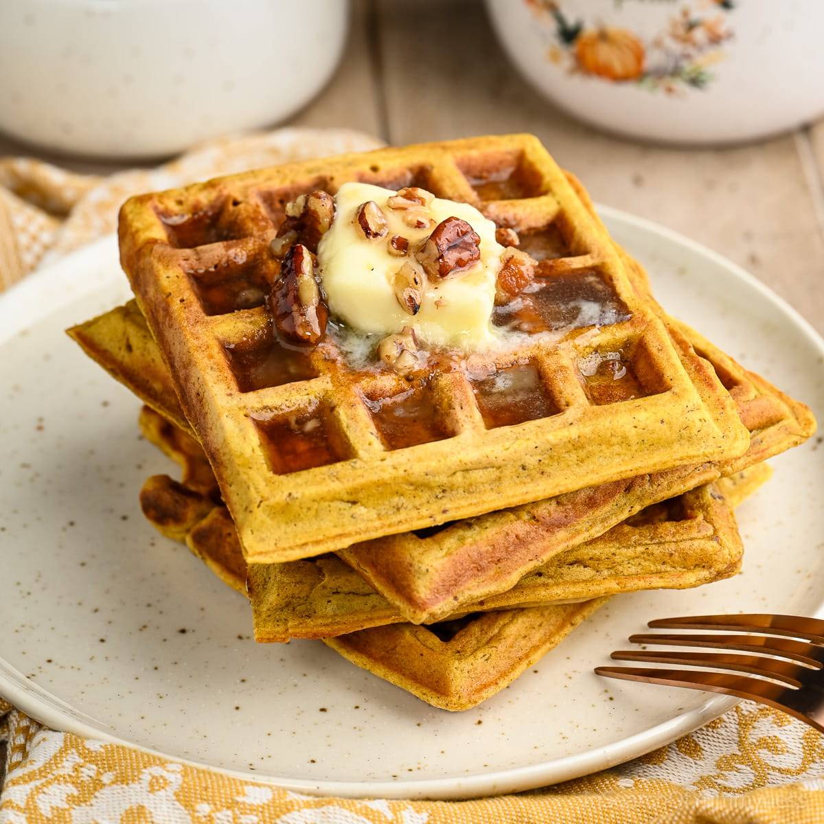 A stack of four golden pecan pumpkin waffles topped with a pat of butter and warm maple pecan syrup, on a beige plate.