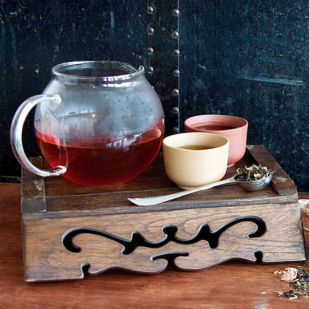 A clear teapot next to two small cups and a tea spoon.