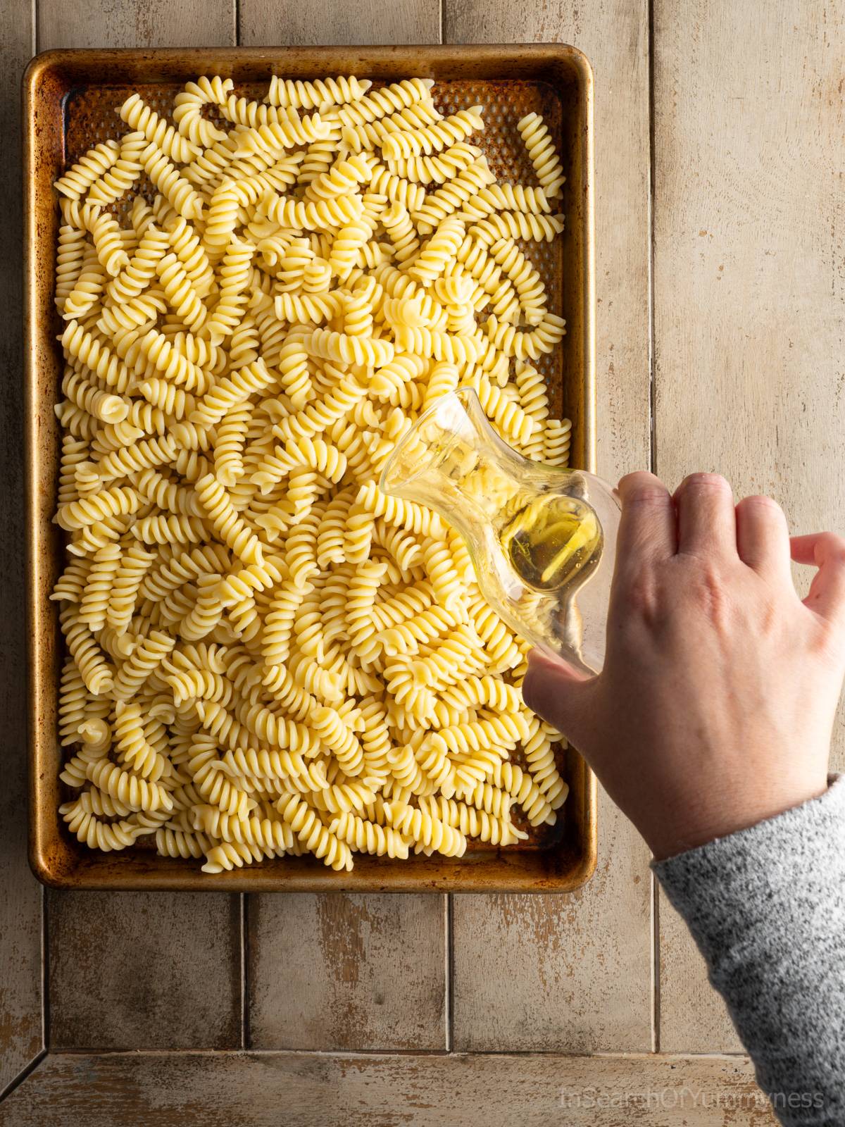 Someone adding oil to freshly cooked pasta cooling on a sheet pan.
