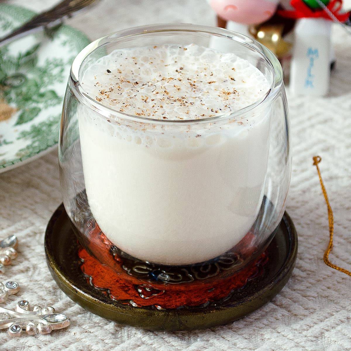 A clear glass of frothy eggless eggnog on a fancy coaster.