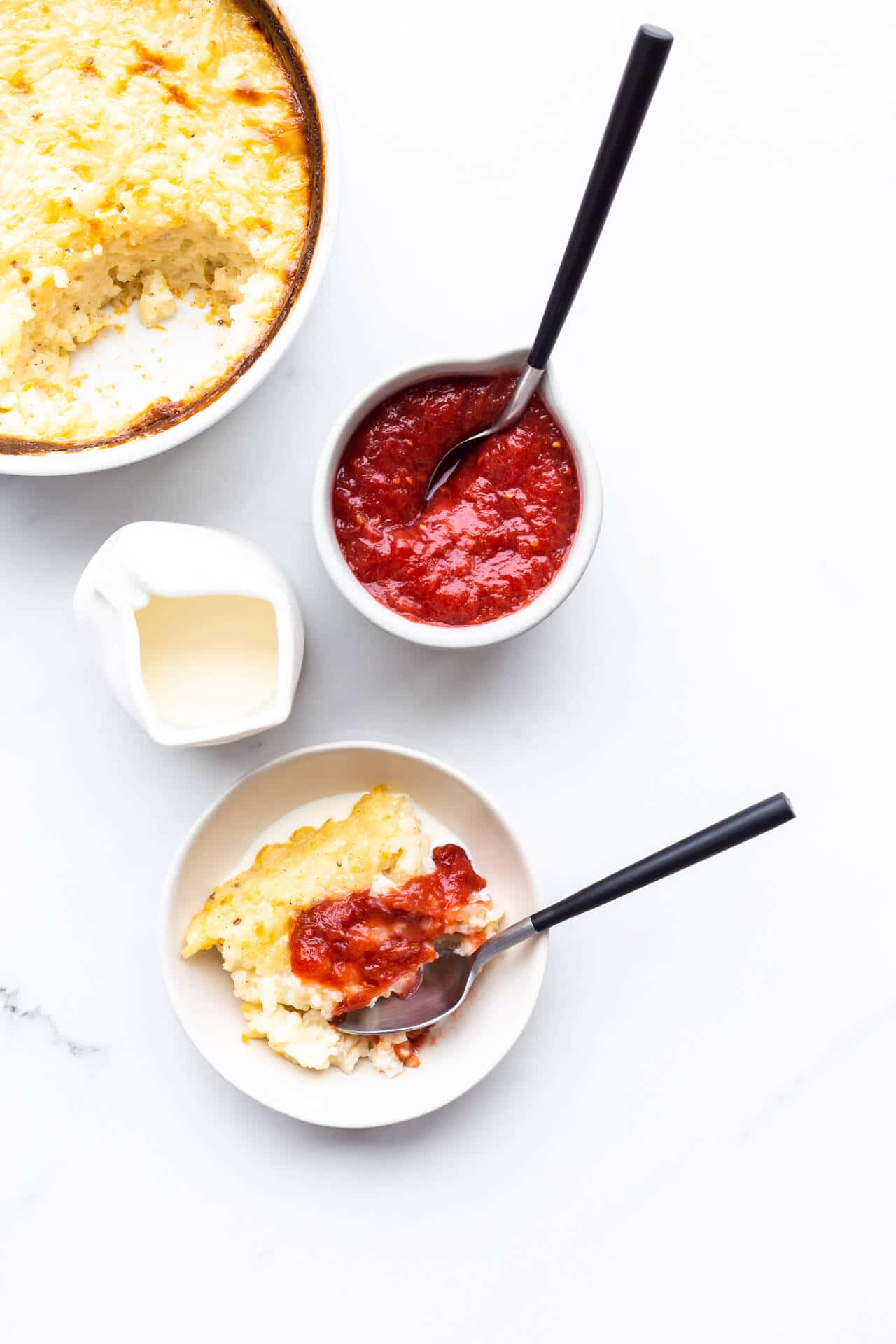 A bowl of baked rice pudding topped with rhubarb jam, next to a pitcher of cream.