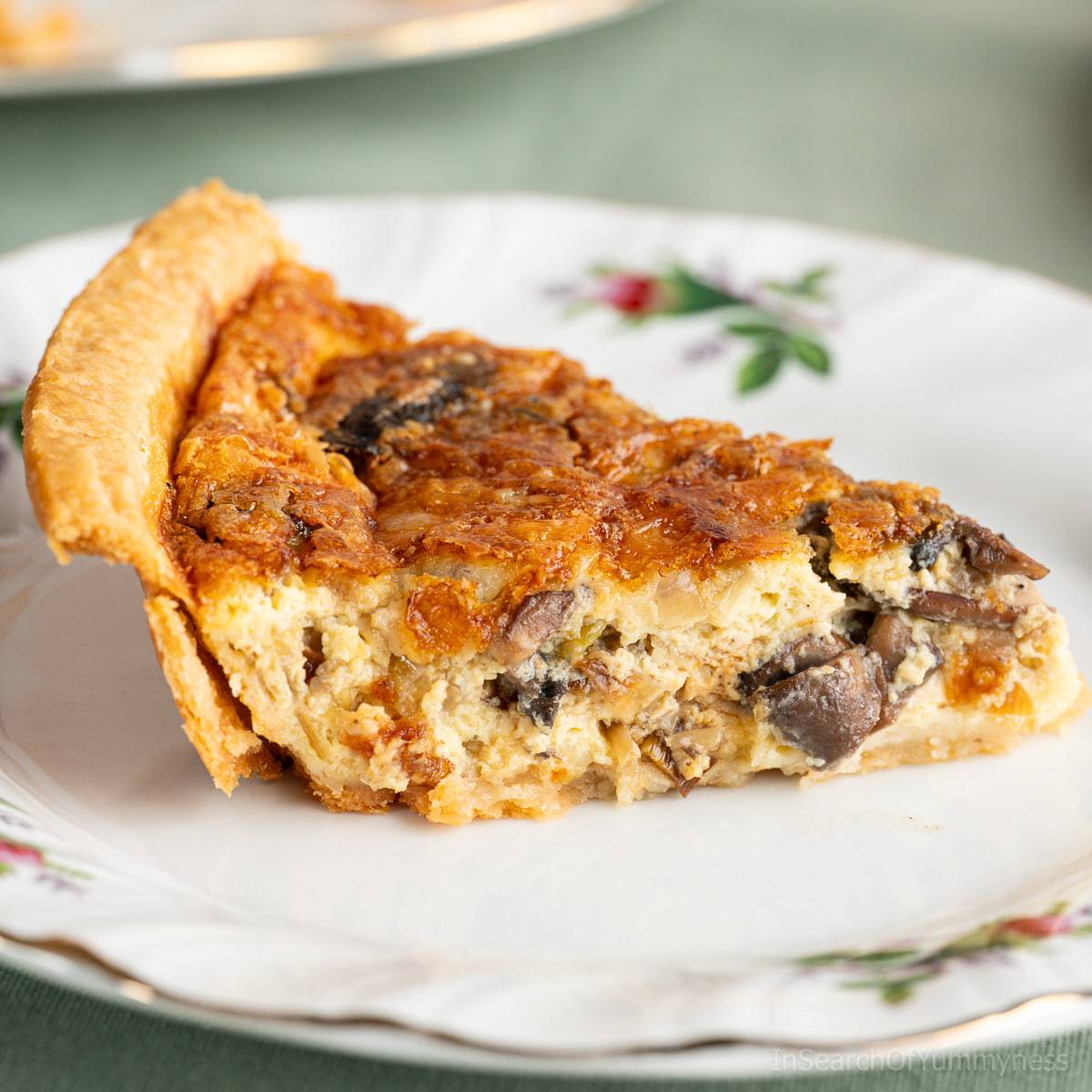A slice of leek and mushroom quiche on a fancy white plate, sitting on a green tablecloth.