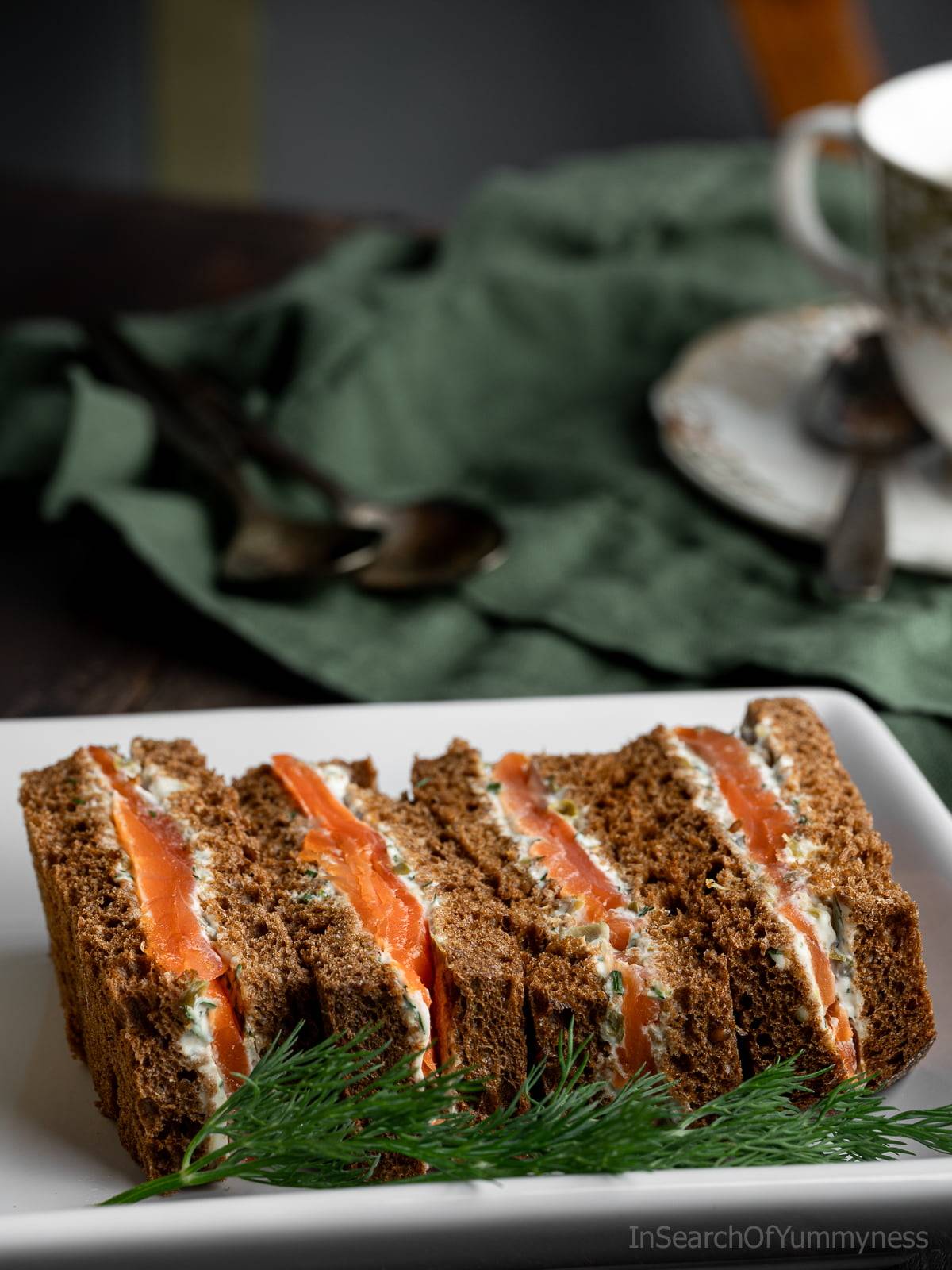 Four smoked salmon tea sandwiches on a white plate next to a green napkin.