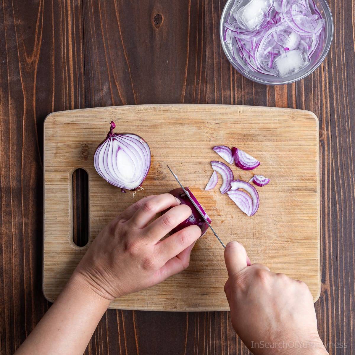 Someone slicing red onions with a knife on a cutting board. There is a bowl of sliced onions sitting in ice water above the board.