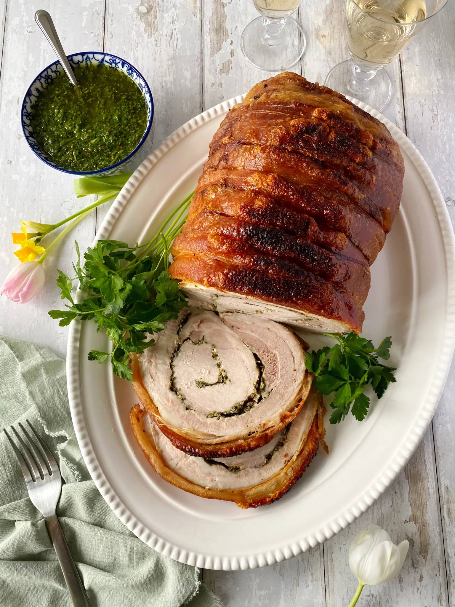 A beautiful porchetta on a cream coloured platter next to a bowl of salsa verde.