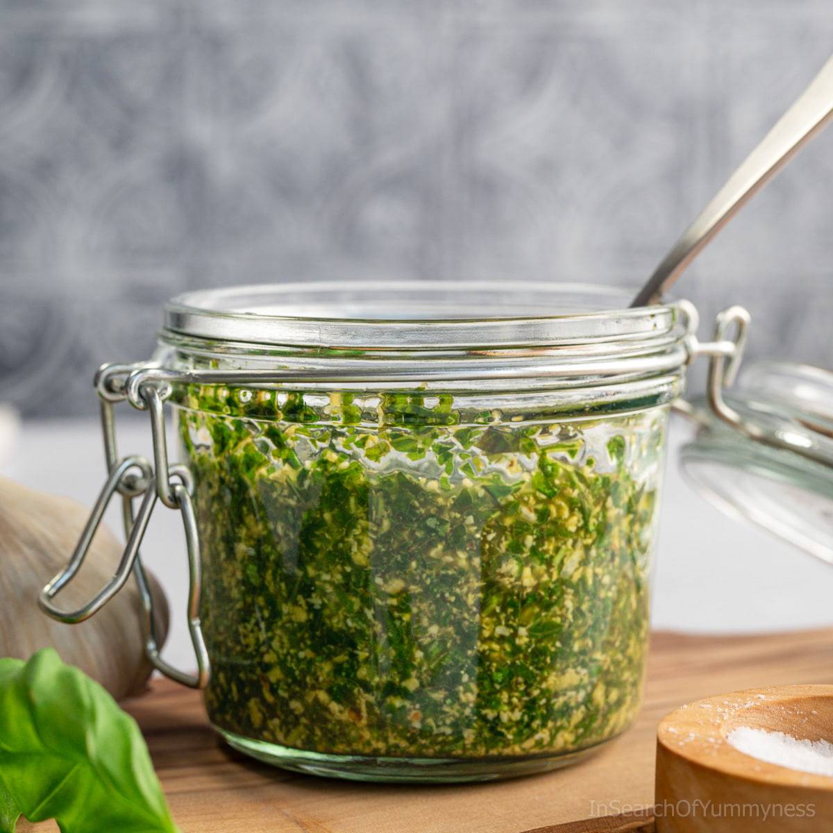 A glass jar filled with vibrant green basil pesto with walnuts, with a spoon sitting in the jar. 
