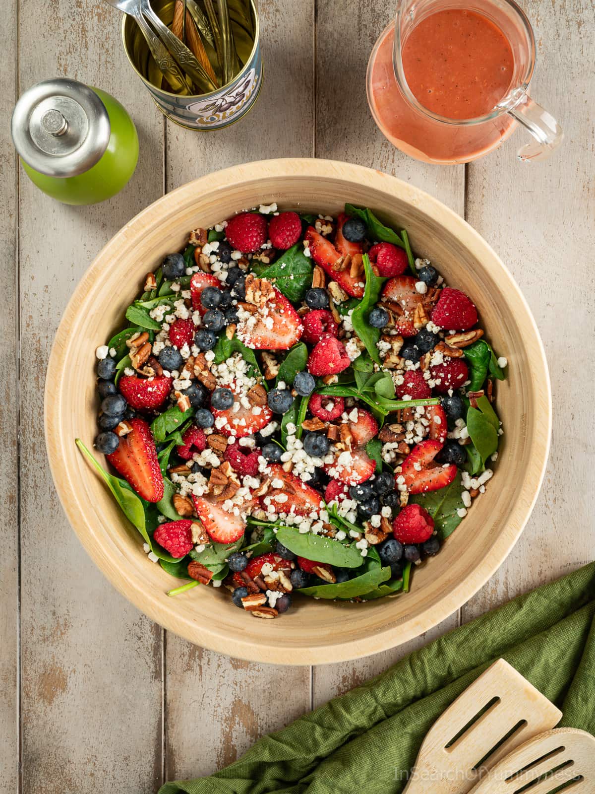 A wooden bowl full of baby spinach, fresh berries, goat cheese crumbles and toasted pecans, next to a glass jug of raspberry vinaigrette, a pepper mill, a can of forks and wooden salad servers.
