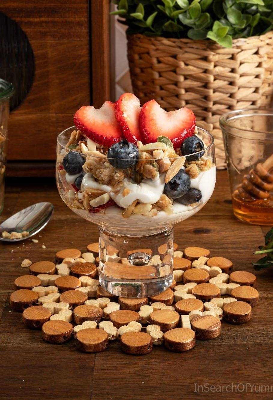 A wide parfait glass with yogurt, granola, almonds and berries, on a table next to a bread box.