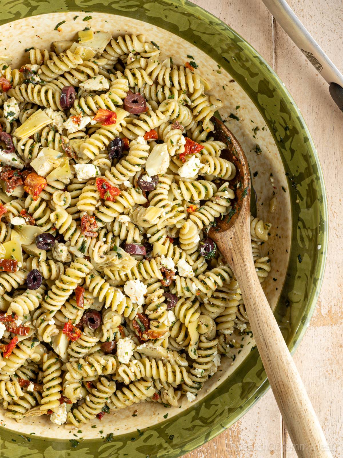 A large ceramic bowl full of pesto pasta salad with feta, olives, sun-dried tomatoes, and marinated artichokes.