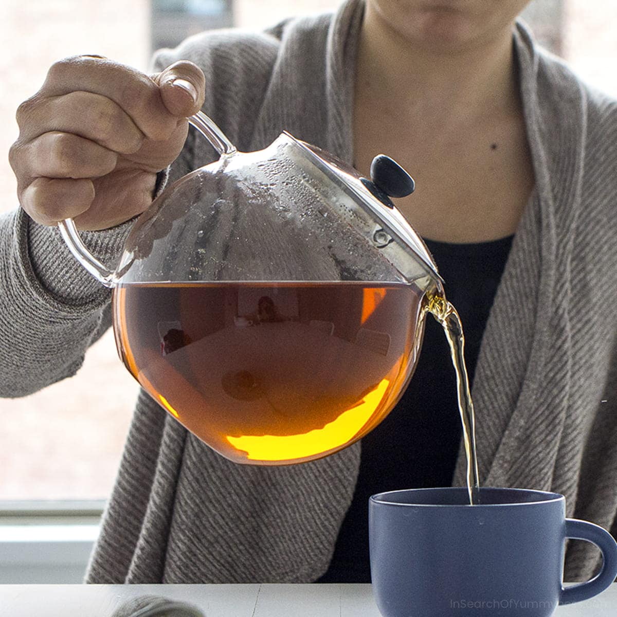 Someone pouring tea from a clear teapot into a blue mug.