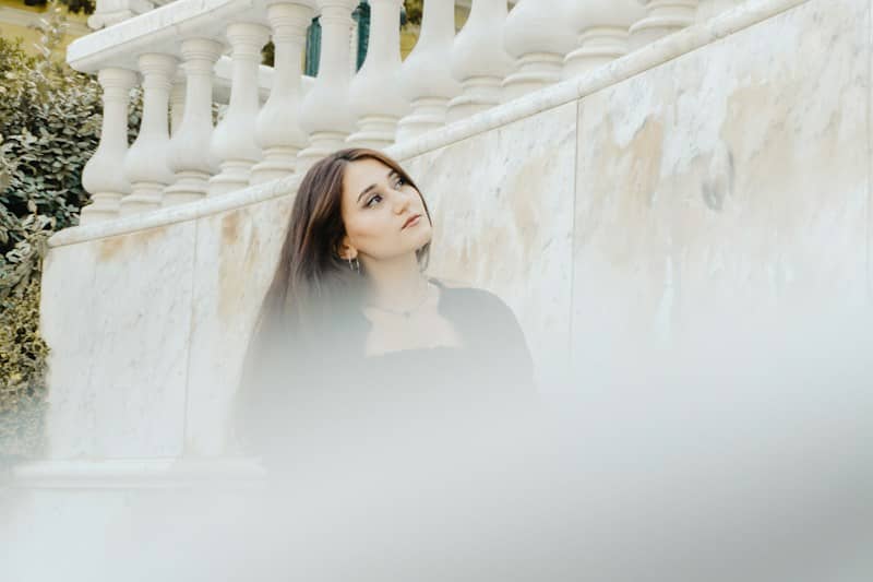 A woman with long dark hair leaning against a marble wall with balustrades