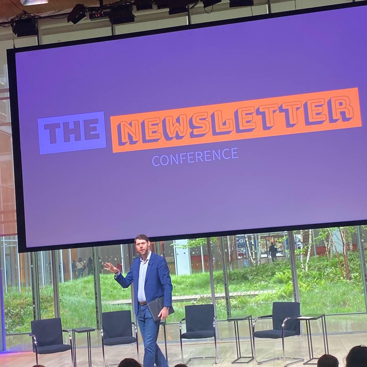 Dan Oshinsky on stage in front of a screen with The Newsletter Projected on it. Behind the stage and screen is a courtyard with trees and plants.