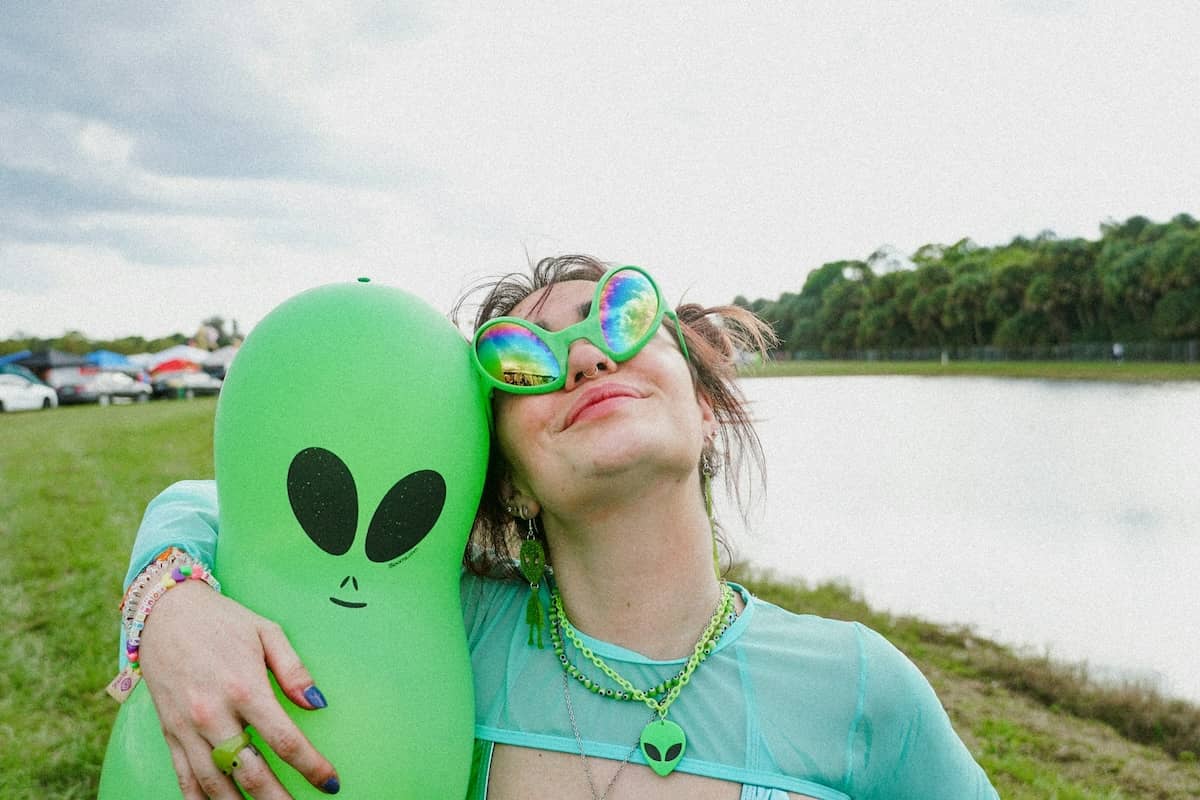 A woman stands in front of trees and a lake. She is wearing shiny green sunglasses and looking up in the sky while holding an inflated plastic alien. As one does.