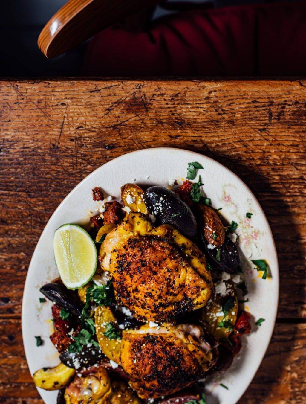 Roasted chicken thighs with crispy, spice-crusted skin served with potatoes, chorizo, crumbled feta, fresh herbs, and a lime wedge on a white platter.