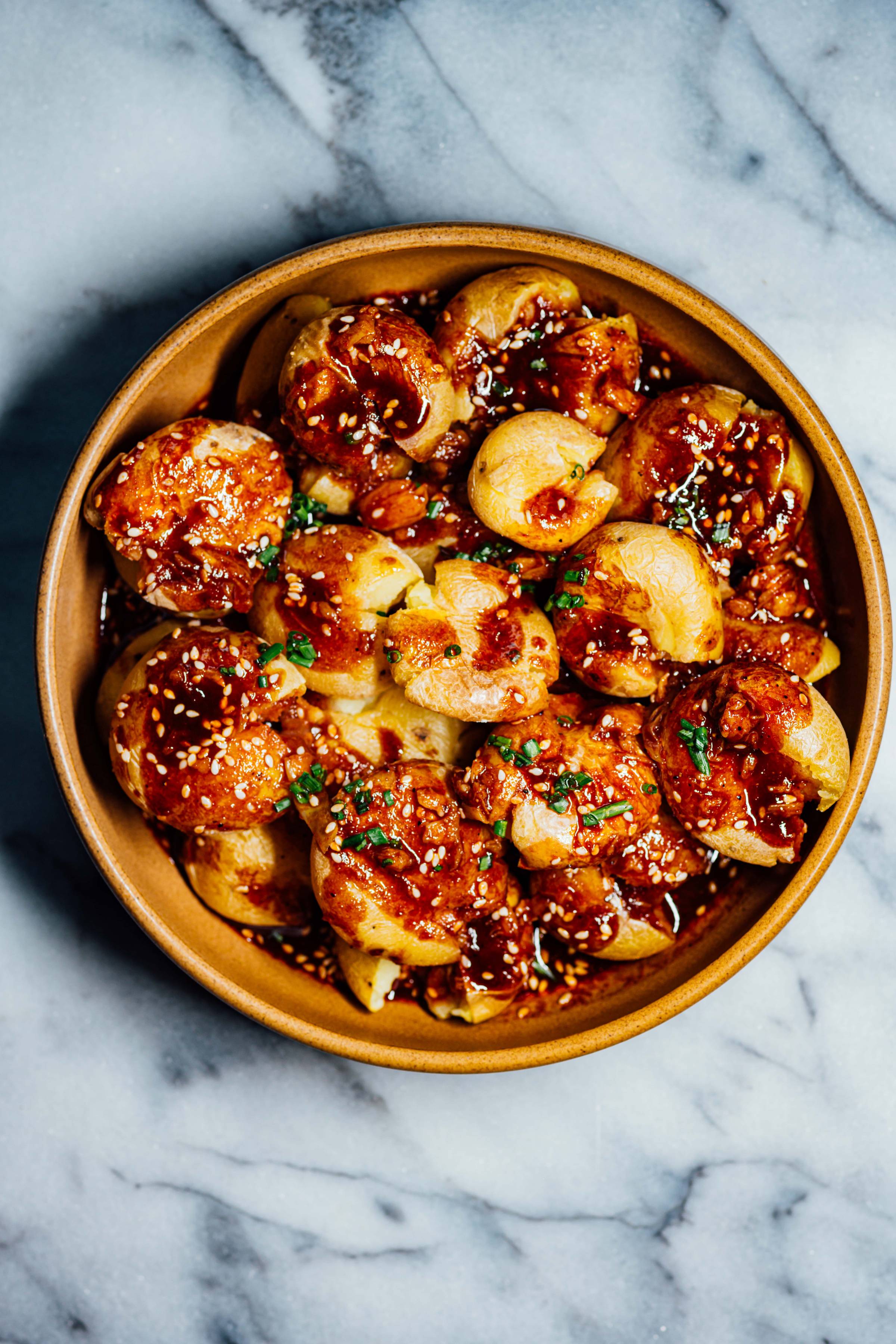Salt-baked new potatoes coated in a glossy gochujang sauce, topped with sesame seeds and chopped chives in a shallow brown bowl.