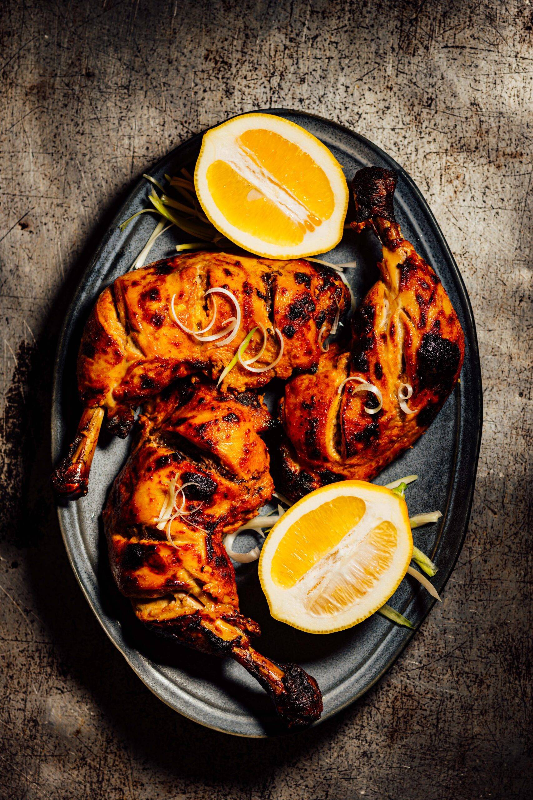Tandoori chicken with a deeply charred, red-orange crust, served on a dark platter with fresh lemon halves and thinly sliced scallions.