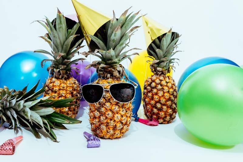 A bunch of pineapples wearing party hats and balloons on a table. One pineapple is wearing sunglasses.