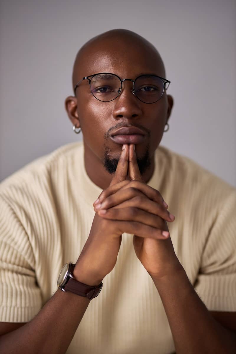 A bald black man with glasses, hands clasped, looking forward.