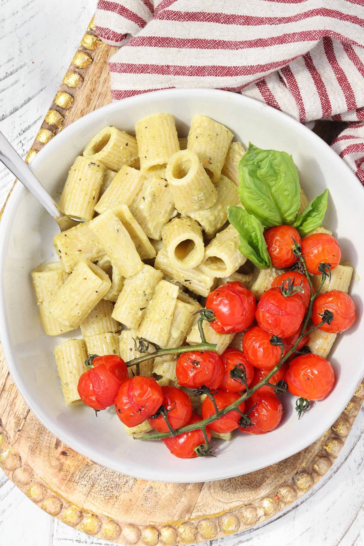 Bowl of rigatoni pasta coated in creamy pesto sauce, topped with roasted cherry tomatoes on the vine and fresh basil leaves.