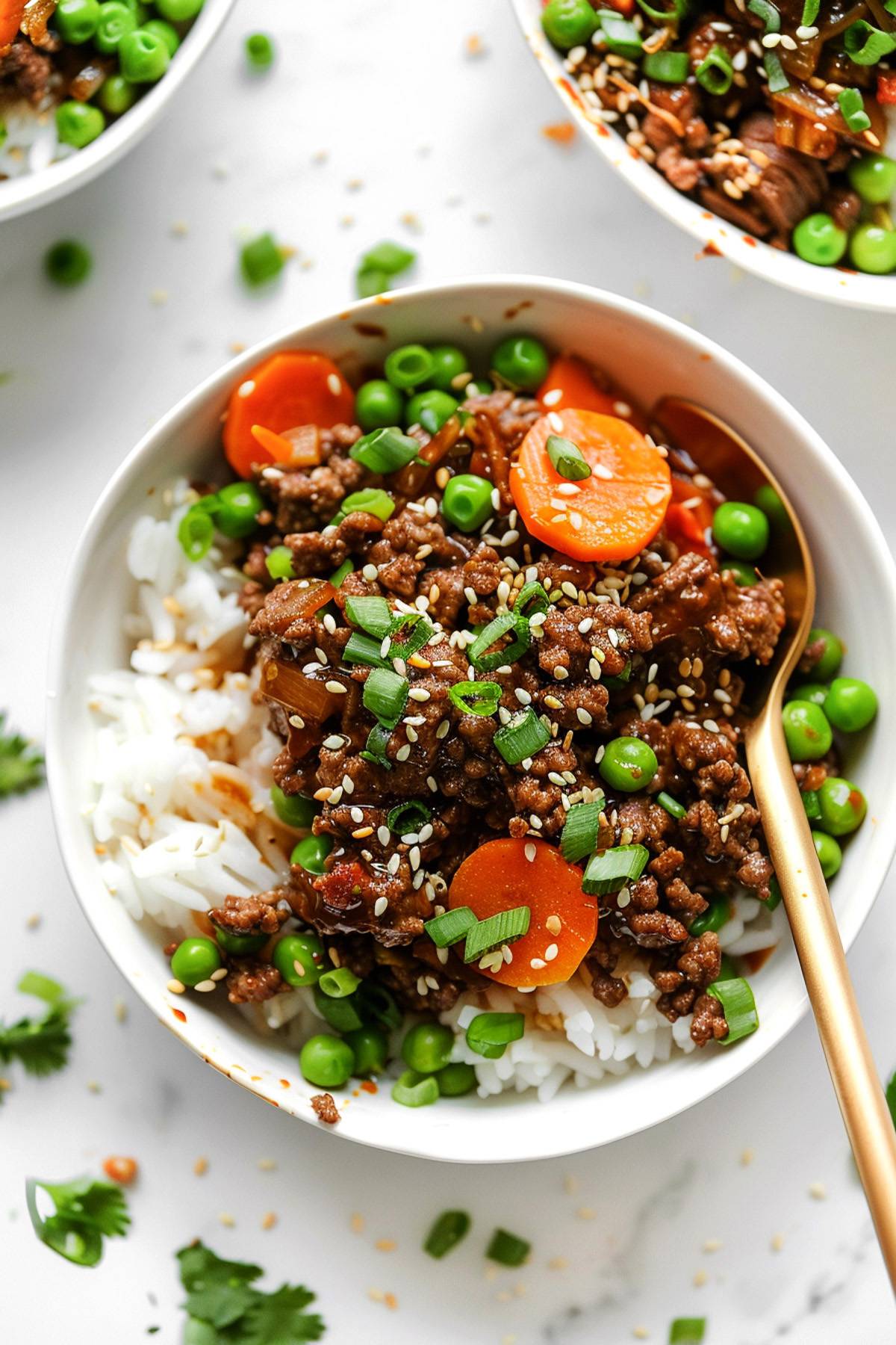 A white bowl filled with Korean beef bowls made of ground beef, carrots, and peas served over white rice, topped with sesame seeds and sliced green onions, with a gold spoon on the side.