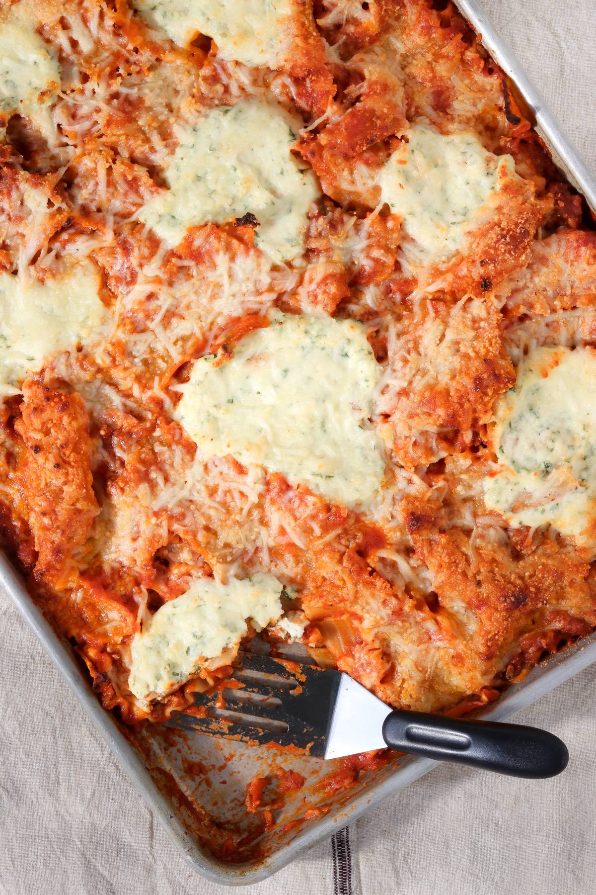 Sheet pan lasagna with broken noodles, marinara sauce, melted mozzarella, and dollops of ricotta, partially sliced with a spatula.