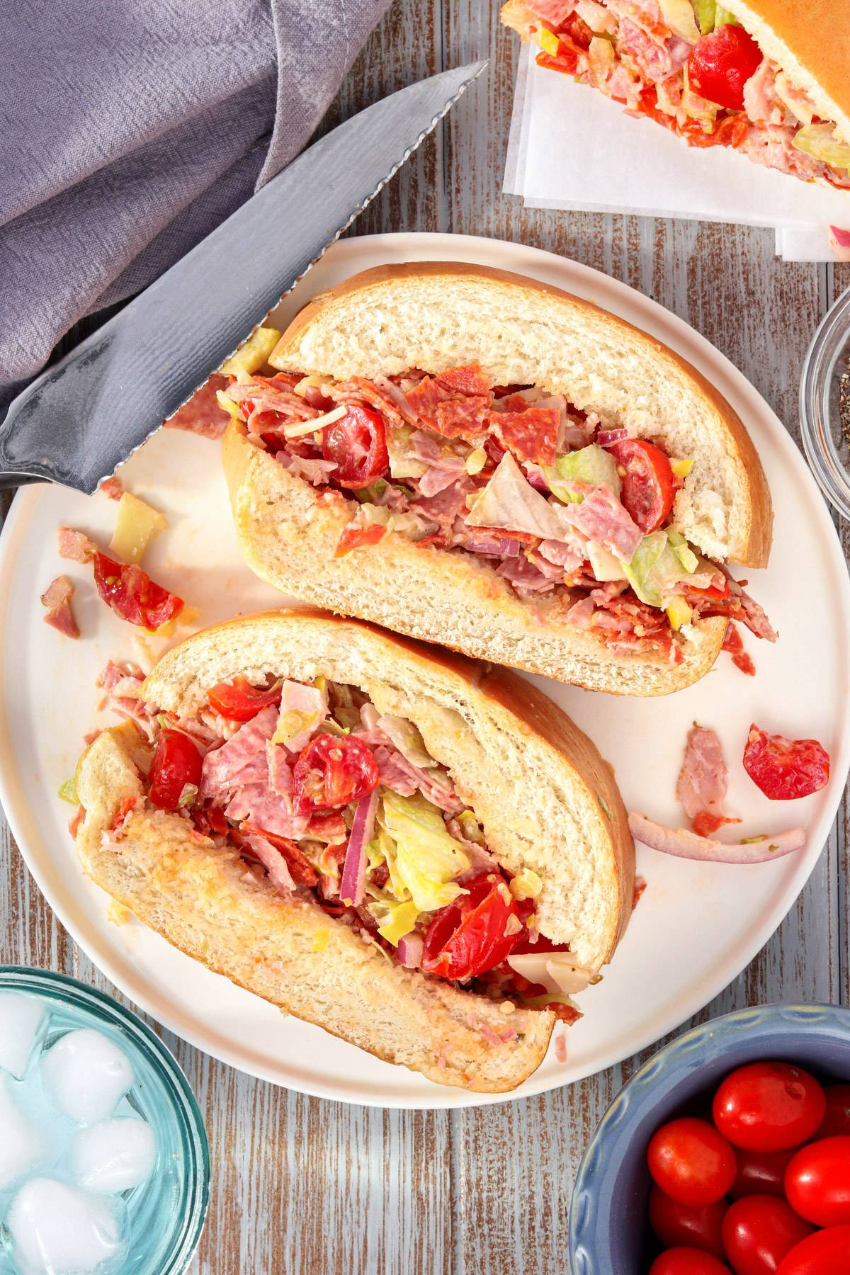 Chopped Italian picnic sandwich cut in half on a white plate with cherry tomatoes and a serrated knife nearby.