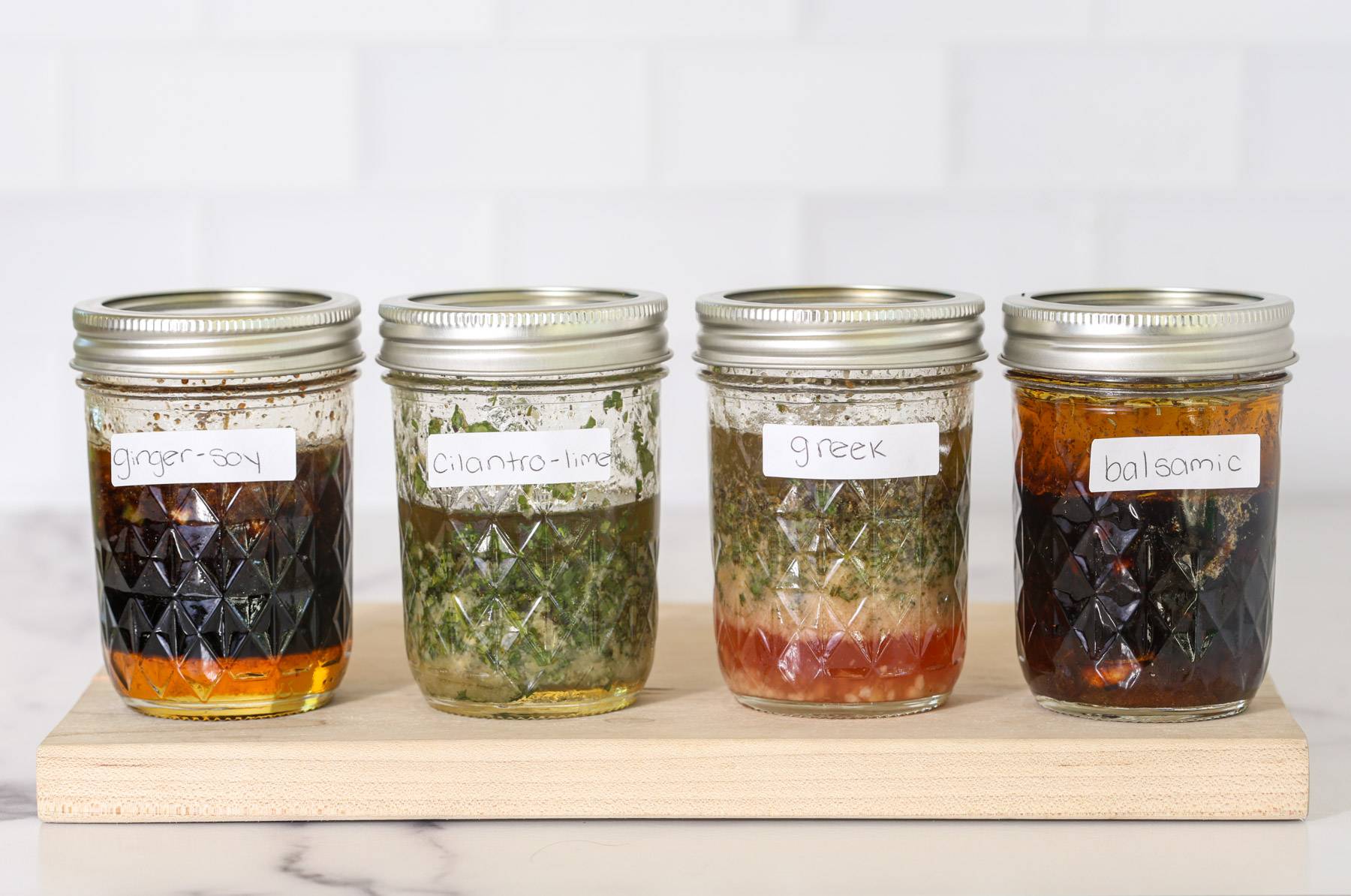 Four small mason jars of chicken marinade lined up on a wooden board, each labeled by hand: ginger-soy, cilantro-lime, greek, and balsamic.
